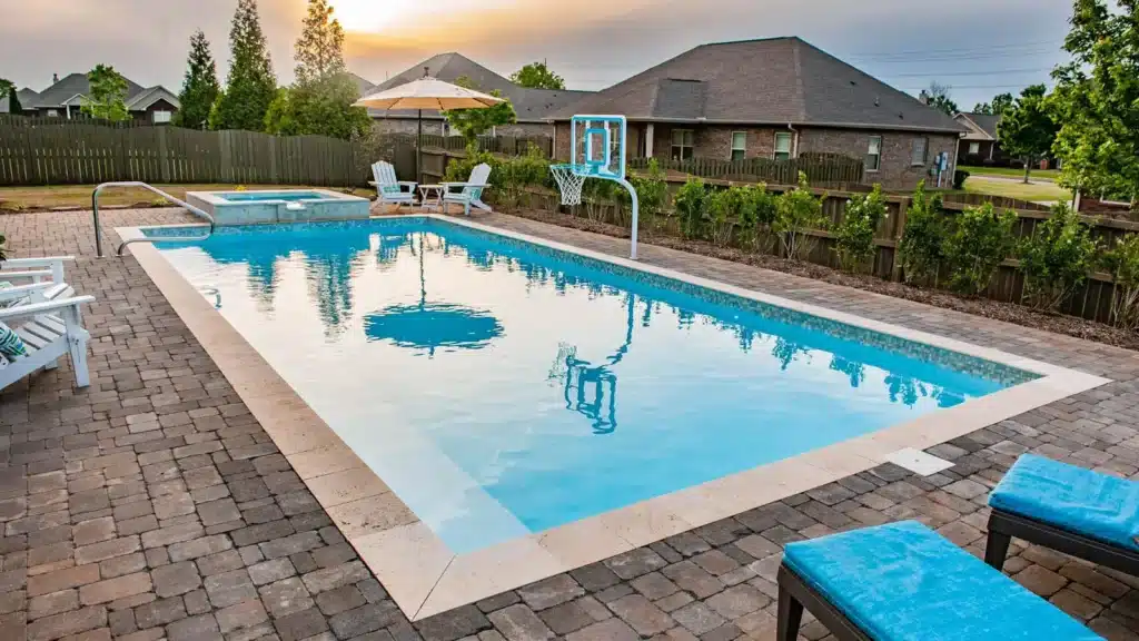 An outdoor swimming pool with clear blue water, surrounded by paver stones. There is a jacuzzi attached, a basketball hoop, and white lounge chairs. Trees and houses are visible in the background under a sunset sky.