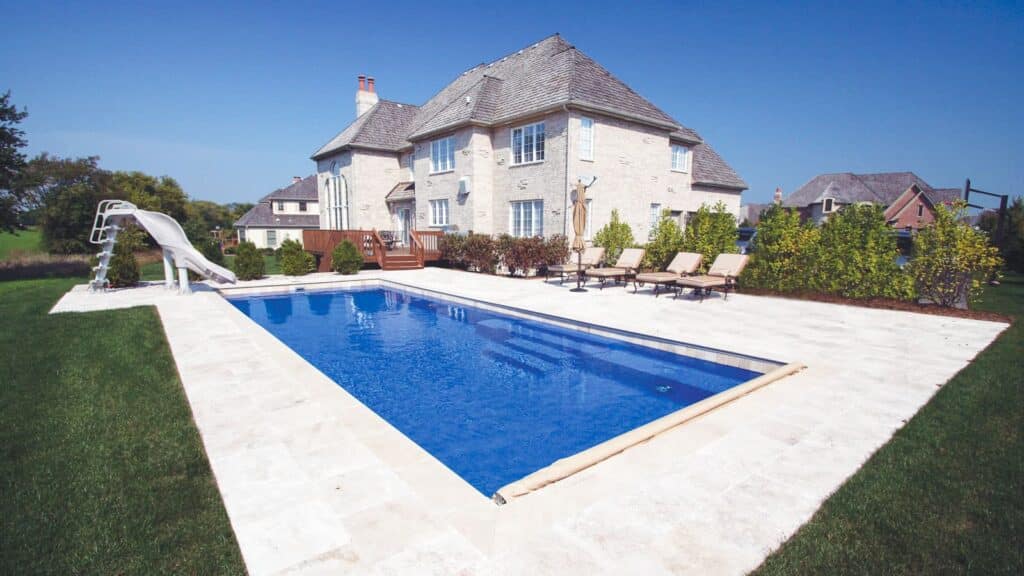 A large backyard featuring a rectangular swimming pool with a slide, surrounded by lounge chairs and well-maintained greenery, next to a spacious stone house under a clear blue sky.