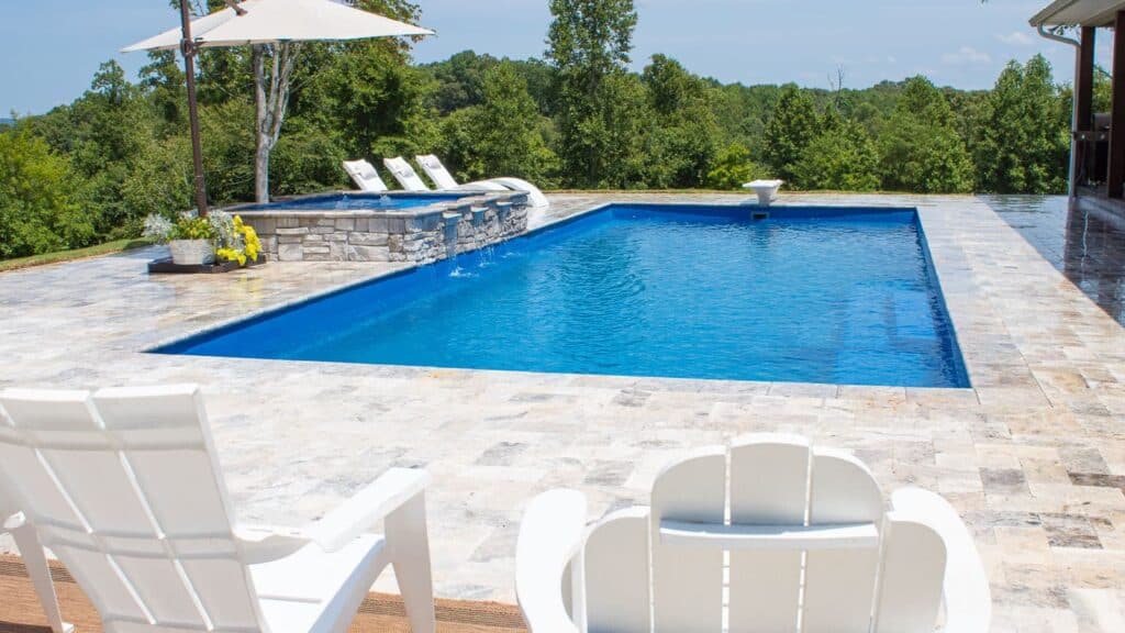 A view of a swimming pool surrounded by a stone deck, with lounge chairs and an umbrella nearby, set against a backdrop of trees and a clear blue sky.
