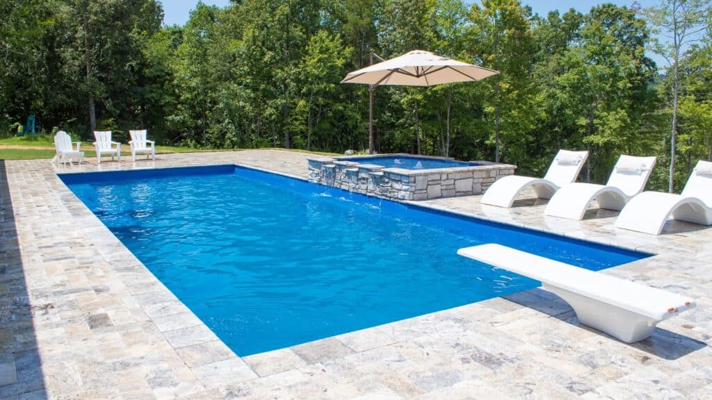 A rectangular in-ground swimming pool with clear blue water, surrounded by stone patio. Wooden lounge chairs and an umbrella are nearby, with greenery in the background.