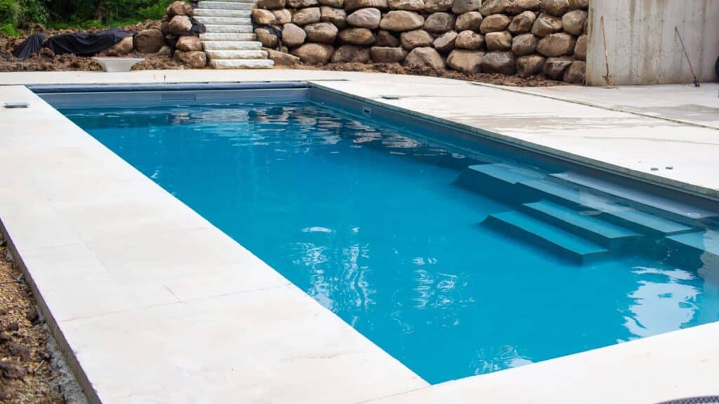 A rectangular swimming pool with clear blue water, surrounded by a concrete patio. Steps lead into the pool, with a stone wall visible in the background. The area is under construction, with bare ground and uninstalled features nearby.