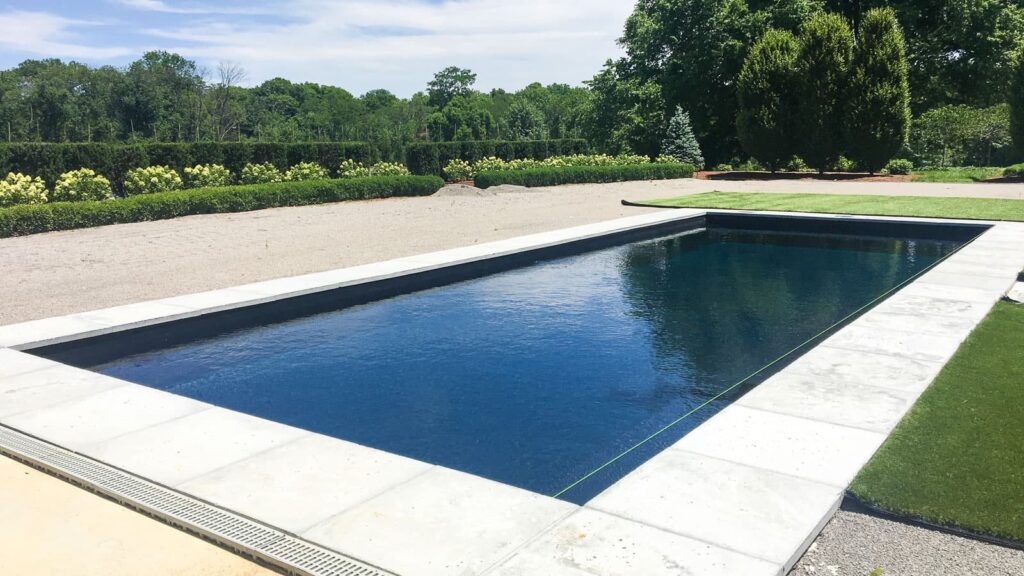 A modern swimming pool with a sleek rectangular design, surrounded by a stone deck and landscaped greenery in the background, under a clear blue sky.