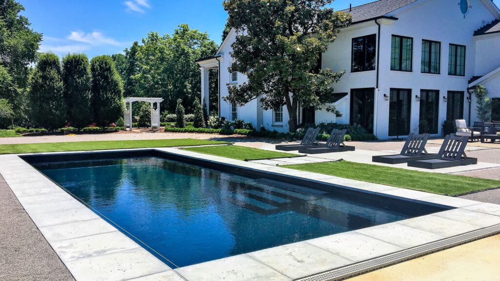 A modern backyard featuring a dark blue rectangular pool with a stone deck, surrounded by lush green grass and trees. Two lounge chairs are placed on the poolside, and a pergola is visible in the background near the house.