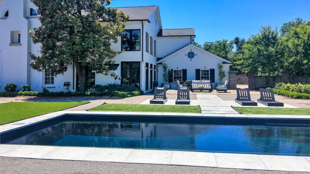 A modern white house with large windows and a sloped roof, featuring a patio area with lounge chairs. A rectangular swimming pool is in the foreground, surrounded by a well-manicured lawn and landscaping.