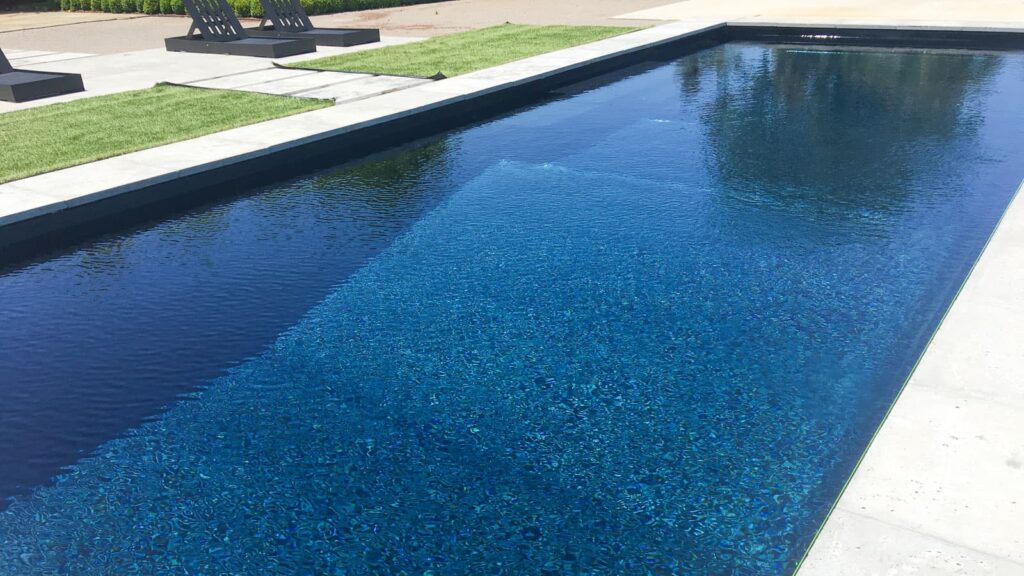 A clear, blue swimming pool surrounded by a modern patio and green grass. Lounge chairs are visible in the background on a sunny day.