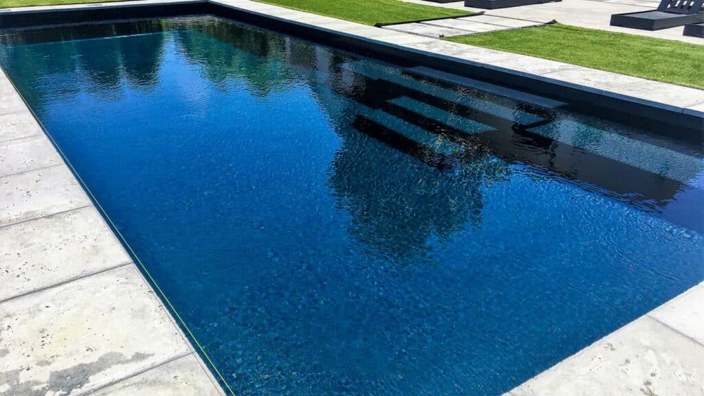 A clear swimming pool reflecting the surrounding landscape, with a sleek, modern design and dark blue water. The pool is bordered by stone tiles and green grass.