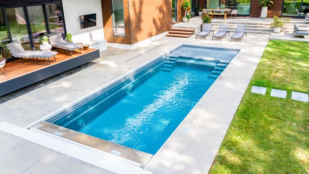 An aerial view of a modern backyard featuring a rectangular swimming pool surrounded by concrete decking. Lounge chairs are positioned nearby, set against a backdrop of a contemporary home with large windows.