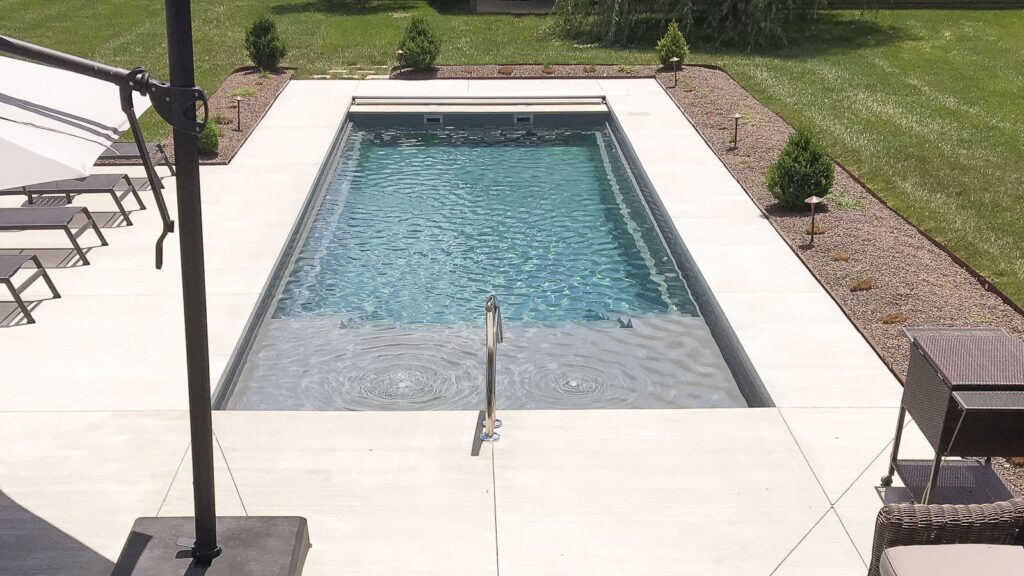An outdoor swimming pool with clear water, surrounded by a concrete deck. Several lounge chairs are positioned nearby, along with grass and small shrubs in the background.