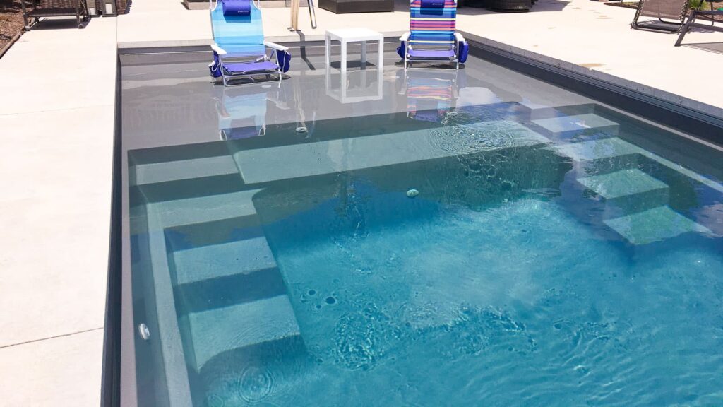 A rectangular swimming pool with steps on one side, featuring clear blue water. Two lounge chairs with striped fabric are positioned near the pool, and a small white table is set between them. The surrounding area includes concrete decking.