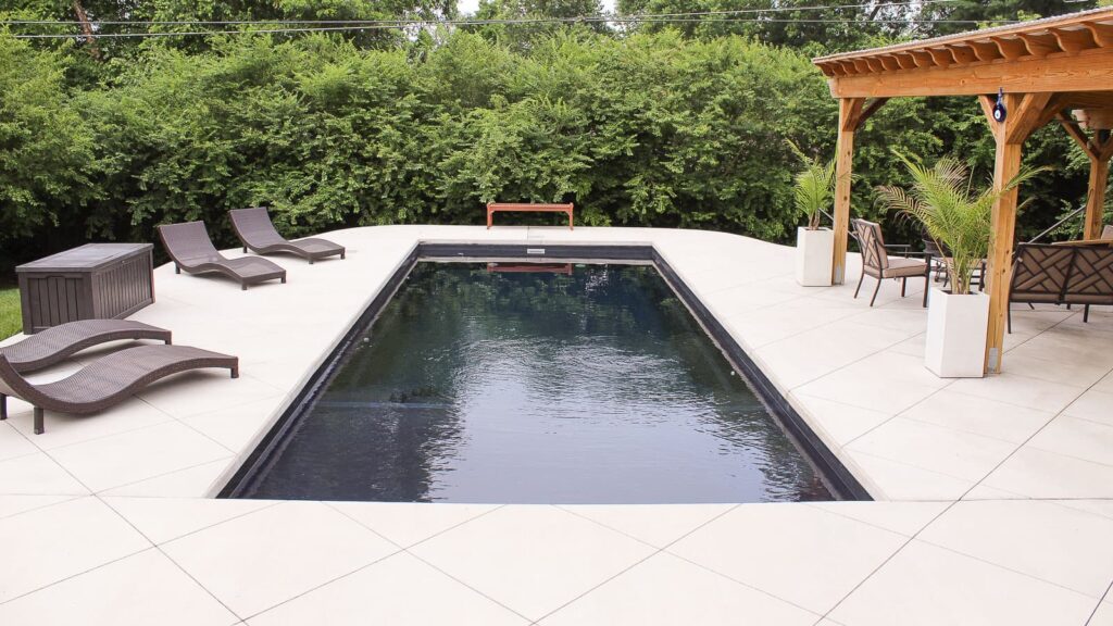 A modern swimming pool surrounded by a tiled deck, featuring two lounge chairs and a seating area with a wooden pergola. Lush green bushes are visible in the background.