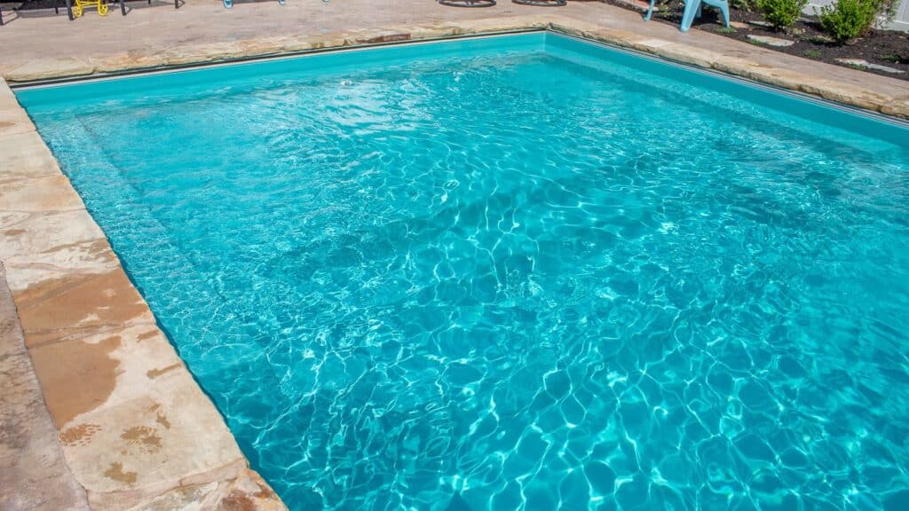 A clear swimming pool with sparkling blue water, surrounded by a stone border and greenery in the background.