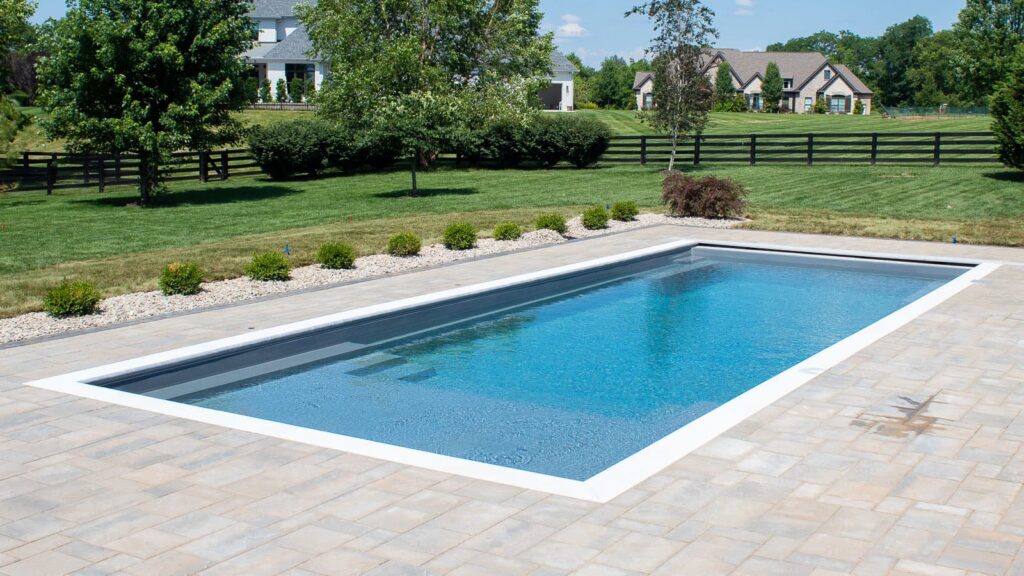 A rectangular swimming pool with clear blue water surrounded by a stone patio, grass, and shrubs in a landscaped yard, with houses visible in the background.