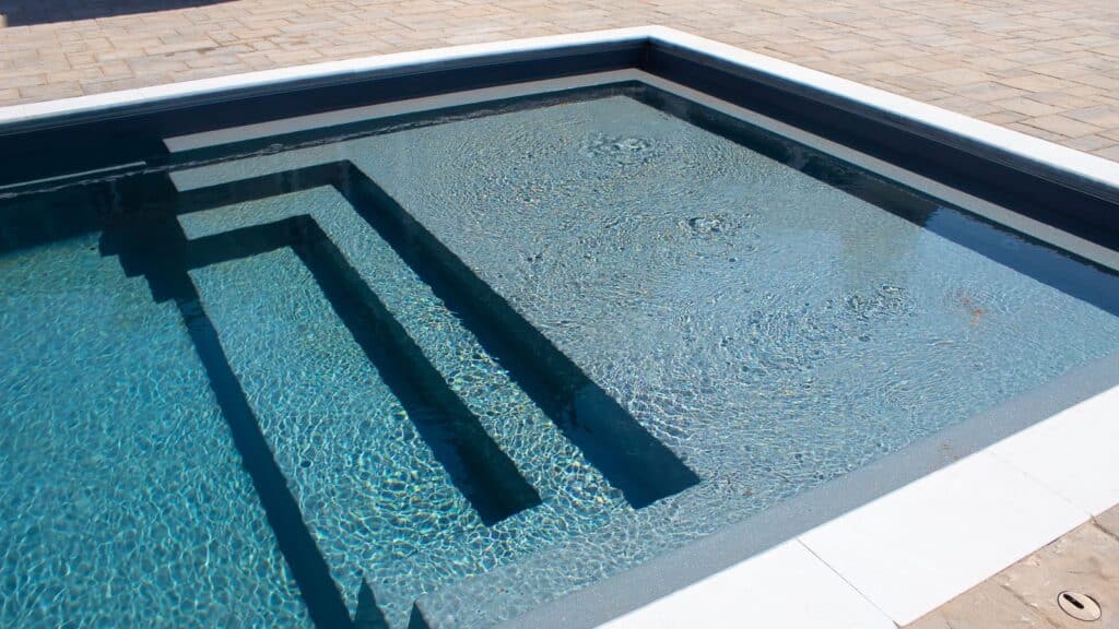 A rectangular swimming pool with clear blue water, featuring steps leading into the water. The pool is surrounded by a light-colored stone deck.