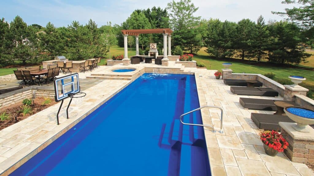 A backyard scene featuring a rectangular swimming pool with a diving board, surrounded by stone patio and landscaped garden. In the background, there are seating areas and a pergola.