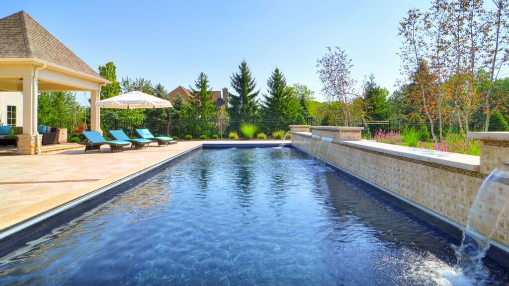 An outdoor swimming pool with clear blue water surrounded by a stone patio. Lounge chairs are placed beside the pool, and greenery and flowers are visible in the background under a clear blue sky.
