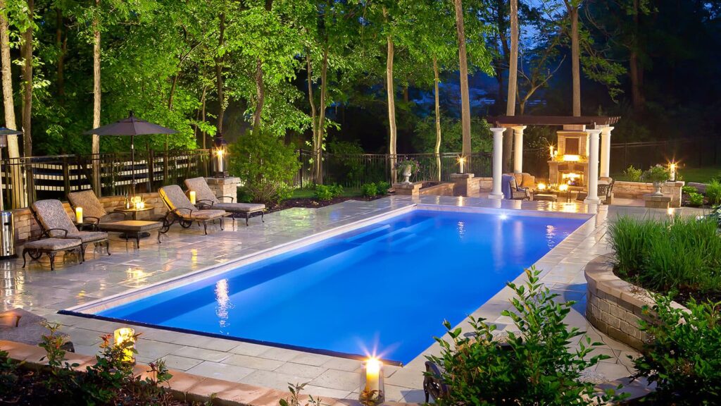 A serene backyard pool area illuminated at dusk, featuring a long blue swimming pool surrounded by lounge chairs, stonework, and landscaped greenery. A pergola with lighting is visible, creating a relaxing atmosphere.