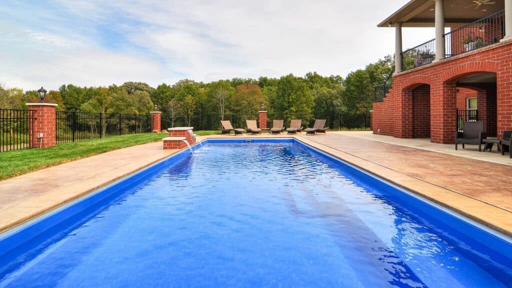 A clear blue swimming pool surrounded by a stone deck, lounge chairs, and a grassy area. Trees are visible in the background under a cloudy sky.
