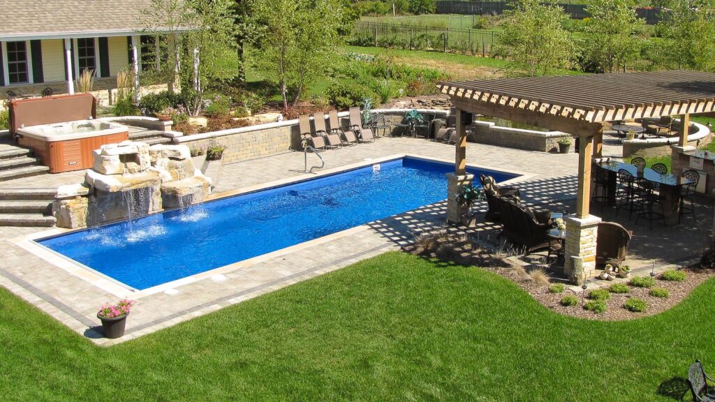 An outdoor pool area featuring a rectangular blue swimming pool with a waterfall, surrounded by a stone patio, lounge chairs, and a pergola. Green grass and landscaped trees are visible in the background.