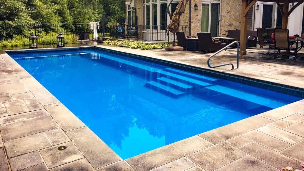 A clear blue swimming pool with steps leading into the water, surrounded by a patio featuring outdoor furniture and a residential building in the background.