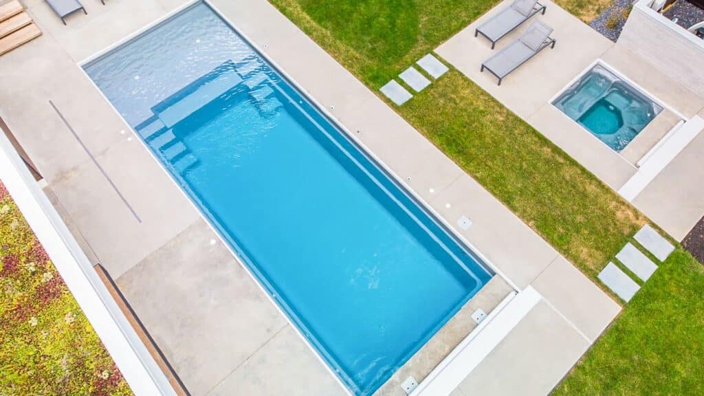 An overhead view of a modern swimming pool surrounded by concrete and grass. Adjacent, a hot tub and two lounge chairs are visible on a landscaped area.