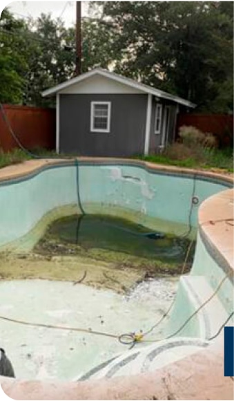 An empty swimming pool with a cracked surface and stagnant water at the bottom, surrounded by overgrown grass. A small gray shed is visible in the background.